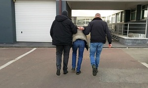 Teren komendy policji, nieumundurowani policjanci prowadzą zatrzymanego mężczyznę.