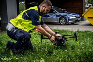 umundurowany policjant trzyma w rękach drona stojącego na trawie, w tle oznakowany radiowóz
