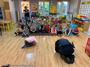 Dzieci w przedszkolu patrzą na inne dziecko i policjanta, który pokazuje pozycje na żółwika.