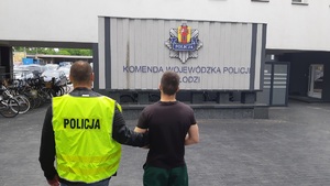 Policjant w kamizelce odblaskowej z napisem policja prowadzi na dziedzińcu komendy wojewódzkiej zatrzymanego oszusta.