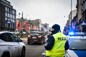 Ulica, policjant stoi tyłem w kamizelce odblaskowej z napisem Policja, po prawej stronie stoi radiowóz na sygnałach świetlnych.