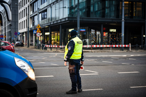 Skrzyżowanie, na rogu skrzyżowania budynki, na drodze stoi policjant w kamizelce odblaskowej z napisem Policja w ręku trzyma tarczę do kierowania ruchem.