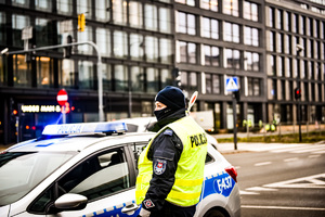 Ulica, radiowóz policyjny, przed nim stoi policjant w kamizelce odblaskowej z napisem Policja w ręku trzyma do góry tarczę do kierowania ruchem.
