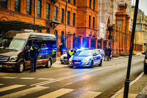 Ulica, po prawej stronie są zaparkowane samochody, na prawym pasie stoi radiowóz na sygnałach świetlnych, przy radiowozie policjant pokazuje palcem kierunek, drugi policjant stoi przy brązowym samochodzie zaparkowanym na poboczu.