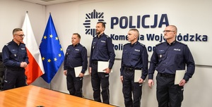 Sala odpraw, po lewej stronie stoi Komendant Wojewódzki Policji w Łodzi na wprost stoją funkcjonariusze policji trzymają w rękach teczki.