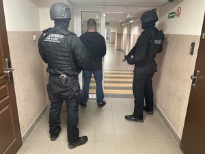 Korytarz zatrzymany stoi tyłem , za nim dwaj policjanci w mundurach, na plechach mają napis Wydział Kryminalny Komendy Wojewódzkiej Policji w Łodzi.