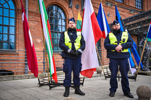Policjanci w mundurach stoją przed flagami