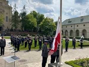 Policjanci stoją na placu w szeregu, flaga Polski wciągana jest na maszt przez poczet flagowy.