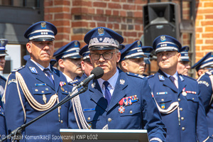 Komendant Wojewódzki Policji w Łodzi przemawia przy mikrofonie, za nim stoi kadra kierownicza.