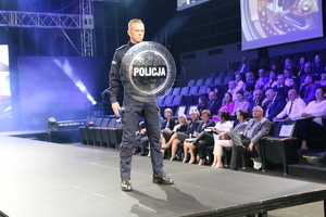 na zdjęciu policjant podczas pokazu mody służb mundurowych prezentuje umundurowanie , stoi na wybiegu, trzymając tarczę z napisem POLICJA, w tle widać publiczność.