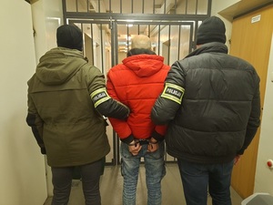 Korytarz, dwa policjanci ubrani po cywilnemu prowadzą zatrzymanego. Przed nimi korytarz z kratą.
