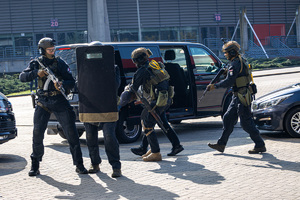 Policjanci z Oddziału Kontrterrorystycznego z bronią długą i tarczami, z tyłu samochody.
