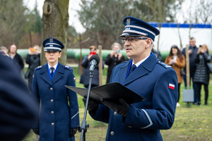 Komendant Wojewódzki Policji w Łodzi czyta przy mikrofonie rotę ślubowania, za nim stoi policjantka.