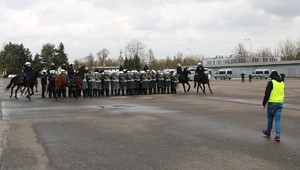 Policjanci z Oddziału Prewencji stoją w tyralierze z tarczami, dookoła policjanci na koniach.