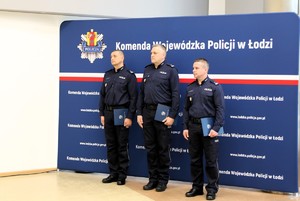 Awansowani policjanci stoją na ściance, w rękach trzymają teczki.