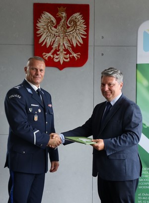 Komendant Wojewódzki Policji w Łodzi i prezes Zarządu Wojewódzkiego Funduszu Ochrony Środowiska i Gospodarki Wodnej stoją na tle godła. Prezes wręcza Komendantowi porozumienie.