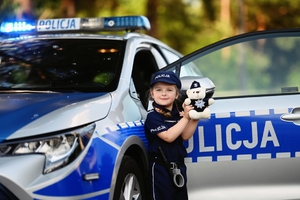 Dziecko w stroju policyjnym stoi przy radiowozie w ręku trzyma misia.