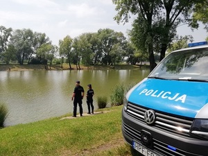 Policyjny radiowóz nad wodą, na brzegu stoi dwóch policjantów.