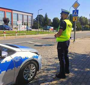 Policjant, obok radiowóz stoi na przeciwko pasów i placówki oświatowej.