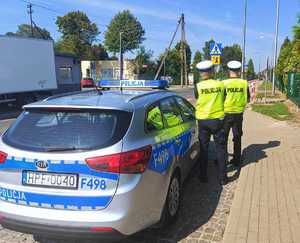 Dwóch policjantów stoi przed przejściem dla pieszych, obok radiowóz.