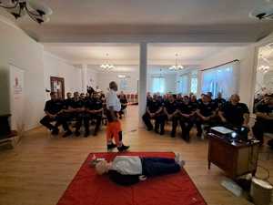 Policjanci siedzą na krzesłach , na podłodze leży manekin, prelegent prowadzi wykład.