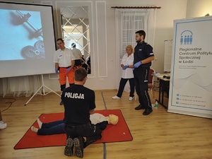 Policjant klęka przed manekinem, drugi policjant stoi, obok prelegent i prelegentka.