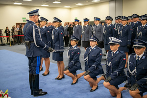 Policjantki klękają na jednym kolanie. Komendant Główny Policji, przykłada do ramienia jednej policjanci szablę, z tyłu stoją w rzędach inni policjanci.