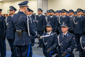 Policjanci klękają na jednym kolanie. Komendant Główny Policji, przykłada do ramienia jednego policjanta szablę, z tyłu stoją w rzędach inni policjanci.