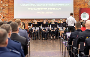 Orkiestra siedzi na krzesłach, w dwóch rzędach siedzą na sali policjanci i strażacy.