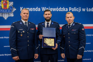 Na ściance stoją Komendant Wojewódzki, wyróżniony policjant który trzyma ryngraf oraz Komendant Powiatowy.