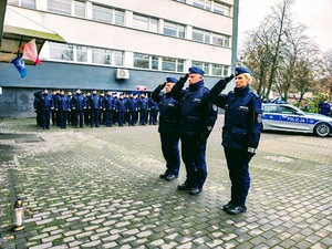 Policjanci stoją w rzędzie. Kierownictwo stoi z tyłu radiowozy policyjne.