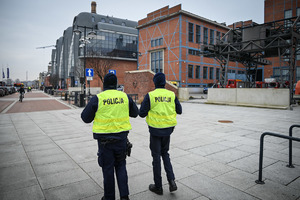 Dwóch policjantów w kamizelkach odblaskowych idą chodnikiem.