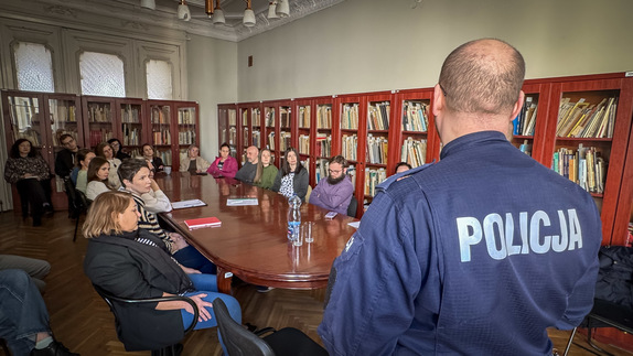 Ludzie siedzą dookoła stołu za nimi regały z książkami z przodu policjant.