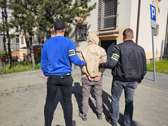 Policjanci kryminalni z opaskami na ręku z napisem policja prowadza zatrzymanego, zatrzymany ma założone kajdanki na rękach.
