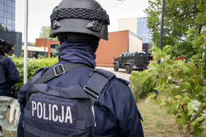 Policjant o d tyłu na pis policja z przodu widać samochód bojowy.