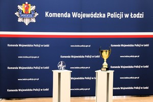 Na tle ścianki Komendy Wojewódzkiej Policji w Łodzi stoi puchar i statuetka.