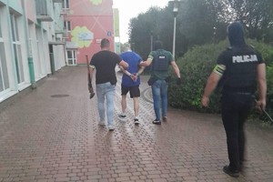 Policjanci idą chodnikiem z zatrzymanym mężczyzną.