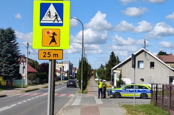 droga, znak drogowy z Agatką, w oddali radiowóz i policjanci ruchu drogowego.