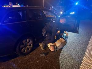 Policjant na ziemi zakłada kajdanki na ręce. Mężczyzna leży na ziemi, obok radiowóz.