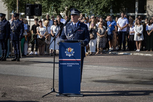 Z Zastępca Komendanta Wojewódzkiego przemawia przy mikrofonie, z tyłu rodziny policjantów.