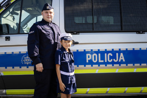 Policjant i dziecko ubrane w strój policjantki przy radiowozie.