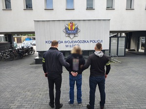 Policjanci prowadza zatrzymanego, zatrzymany ma kajdanki założone na ręce trzymane z tyłu.