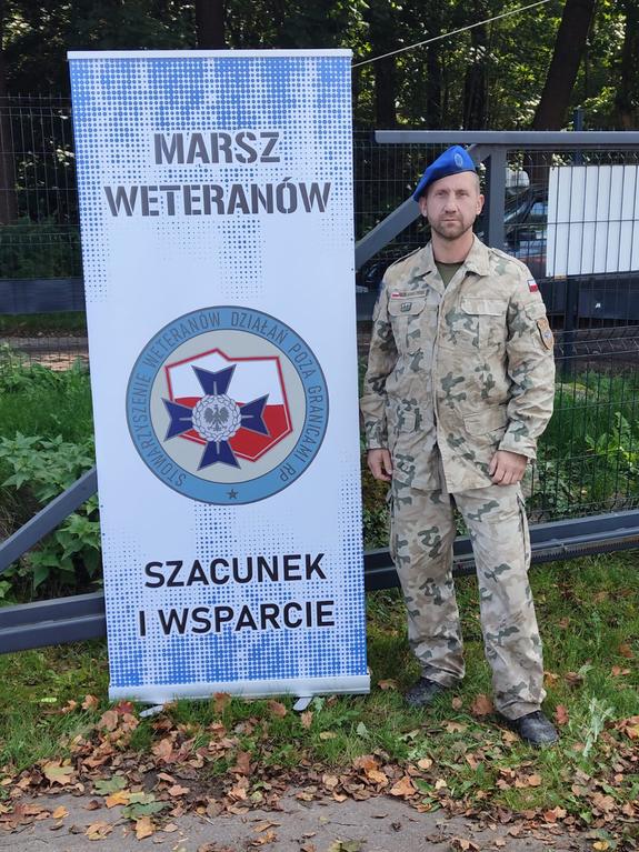 policjant w mundurze koloru moro stoi przy banerze z hasłem Marsz Weteranów, szacunek i wsparcie.