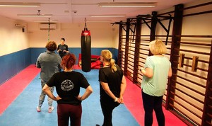 Instruktor prowadzi zajęcia na macie, biorą udział w nich kobiety.