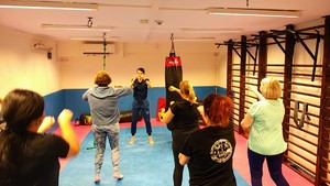 Instruktor prowadzi zajęcia na macie, biorą udział w nich kobiety.