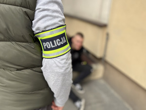 Policjant na rękawie z napisem policja obok siedzi zablurowana postać mężczyzny.