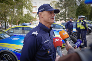 Komendant Wojewódzki Policji w Łodzi w trakcie udzielanego wywiadu. W oddali widać umundurowanych policjantów i oznakowane radiowozy.