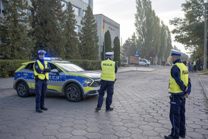 Trzech umundurowanych policjantów stoi przy oznakowanym radiowozie. W tle budynek Starostwa Powiatowego w Zgierzu.