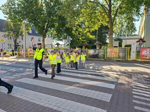 Dzieci w kamizelkach odblaskowych prowadzone przez policjantkę na pasach.