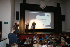 Zdjęcie dwóch prelegentów (jeden w mundurze (przy mównicy) oraz stojący obok-w garniturze) na estradzie na tle wyświetlonej tytułowej strony prezentacji - w dole zdjęcia widoczna część publiczności na widowni
